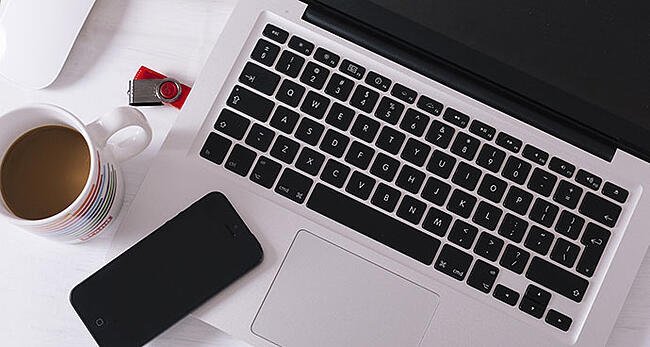 ノート型パソコンとスマートフォンとマグカップに入ったコーヒーが机上に置かれている様子